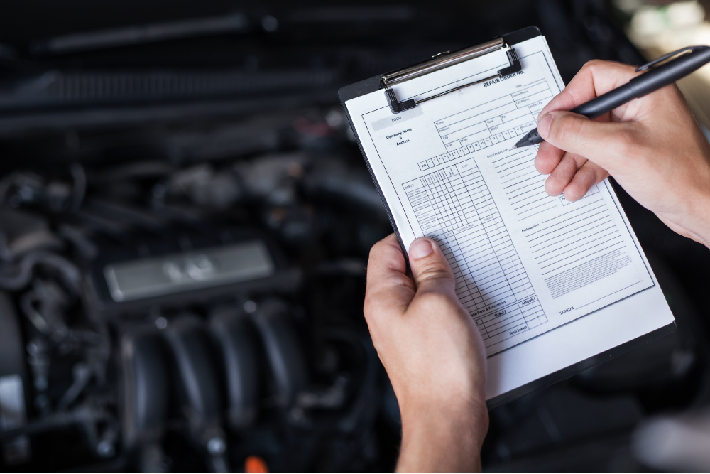 car inspection, Auto Repair & Service, Auto Body Repair & Auto Collision Repair in McAllen, Edinburg & Harlingen, TX. Mechanic filling out a vehicle repair order on a clipboard in front of an open engine bay, documenting service details.
