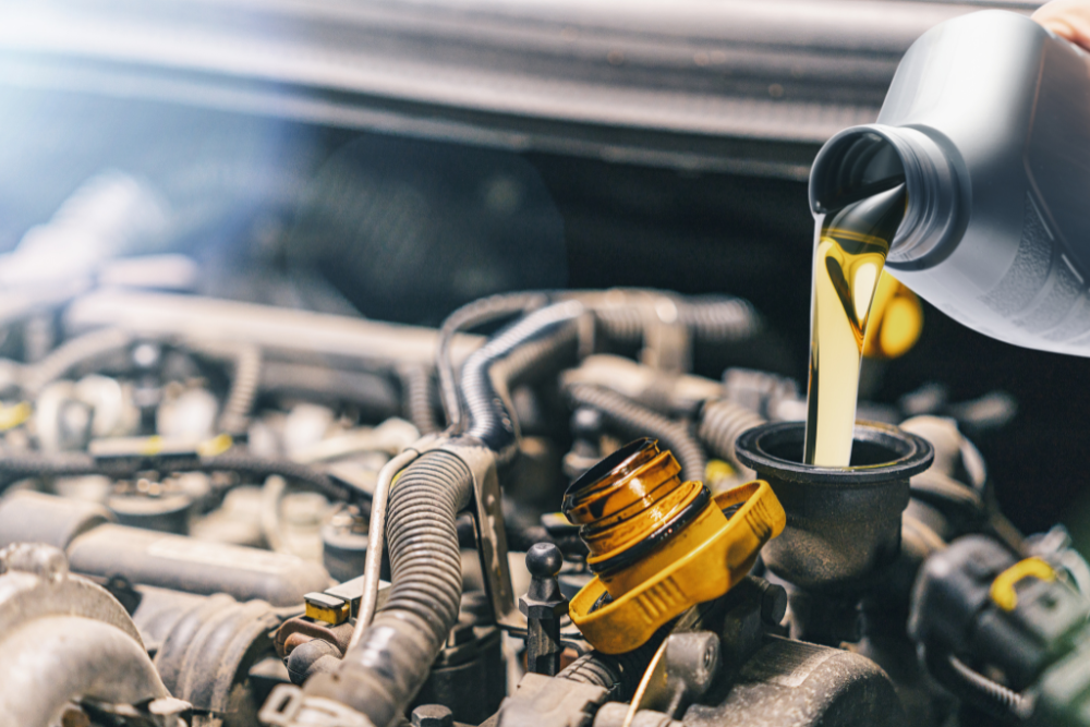 Oil Change or Engine Trouble, Auto Repair & Service, Auto Body Repair & Auto Collision Repair in McAllen, Edinburg & Harlingen, TX. Close-up view of golden motor oil being poured from a bottle into an open engine oil fill port, with various engine components and wiring visible in the background.