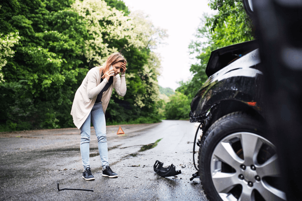 Nissan and Infiniti Certified Collision Repair: Expert Care in Harlingen - Premium Collision - A woman stands next to a damaged car on a deserted road, holding her head and appearing distressed, with car debris scattered on the ground.